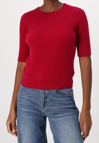 Roter, gerippter Strickpullover mit kurzen Ärmeln und Rundhalsausschnitt, kombiniert mit blauen Jeans aus Denim, die leicht ausgebleicht sind.