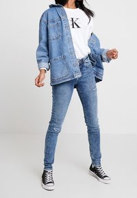 Mujer vestida con chaqueta de mezclilla azul clara, camiseta blanca con logo de Calvin Klein, jeans ajustados desgastados y zapatillas altas negras, de pie contra un fondo blanco.