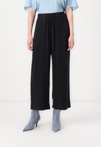 Svart bukser med vid benvidde, elastisk midje, glatt tekstur og en avslappet passform, sammen med blå ankelboots, vist i en nøytral bakgrunn.