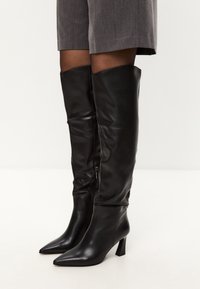 Botas de cuero negro hasta la rodilla con punta afilada y tacón cuadrado. Tienen una textura suave y un cierre lateral para facilitar su uso.