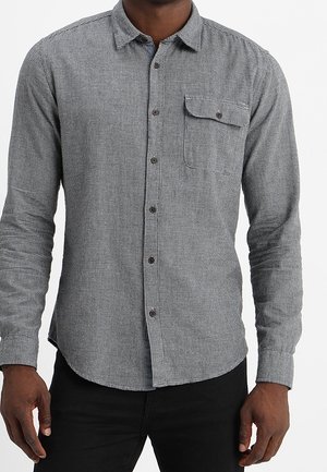 Mężczyzna noszący szarą, długorękawową koszulę zapinaną na guziki z małym wzorem w kratę i zapiętym na guzik kieszonką na piersi, w połączeniu z czarnymi spodniami.