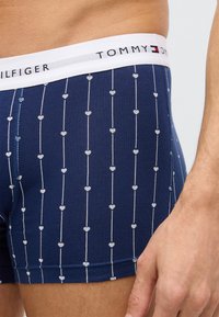 Marineblaue Baumwoll-Boxershorts mit einem weißen Bund, auf dem das "TOMMY HILFIGER"-Logo zu sehen ist, und einem Muster aus kleinen, weißen, vertikalen Herzen.