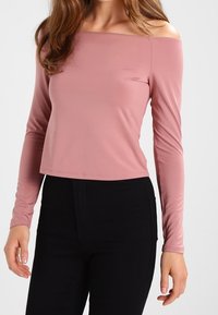 Top s odhalenými rameny a dlouhými rukávy z jemné růžové látky. Vyznačuje se zkráceným střihem a hladkou texturou. Spárováno s černými kalhotami.