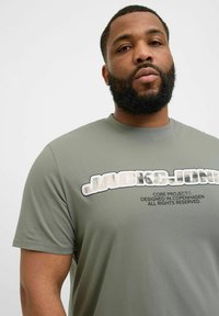 Grøn T-shirt med korte ærmer lavet af blødt stof, der har et dristigt hvidt logo og tekstgrafik på tværs af brystet, med en afslappet pasform.