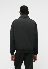 Melna, viegla jakas ar apkakli, elastīgu jostasvietu un aprocēm. Gluda tekstūra ar minimālistisku dizainu, piemērota ikdienas valkāšanai.