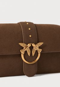 Bruine leren clutch met een gouden ronde hardware detail met twee vogels. Geaccentueerd met gouden studs langs de sluitingstrap.