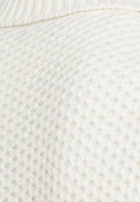 Tessuto a maglia bianco testurizzato con un motivo a nido d'ape e colletto a coste. La superficie appare morbida e spessa, enfatizzando profondità e dimensione.