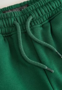 Gröna sweatpants med justerbar dragsko, elastisk midja och mjukt bomullsmaterial. Nära bild visar textur och sömnadsdetaljer.