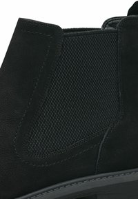 Botín negro hasta el tobillo con acabado de ante, que presenta un panel lateral de malla texturizada, costuras reforzadas y una suela de goma flexible.
