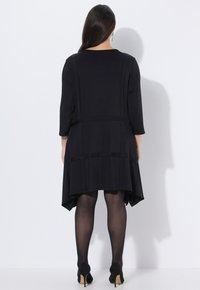 Pitkähiuksinen nainen, jolla on musta epäsymmetrinen mekko, läpikuultavat mustat sukkahousut ja mustat korkokengät, katsoo valkoista seinää.