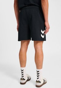 Man in zwarte shorts met witte chevronontwerpen aan de zijkant, witte sokken met zwarte chevrons en witte sneakers met zwarte strepen.