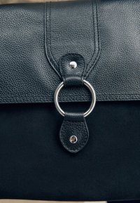 Sac en cuir noir avec rabat texturé, fermoir anneau en métal argenté et deux sangles en cuir maintenues par des clous argentés.