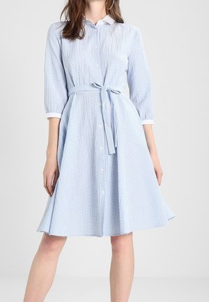Donna che indossa un vestito in gingham bianco e azzurro, lungo fino al ginocchio, con maniche a tre quarti, bottoni frontali, colletto e una cintura legata in vita.