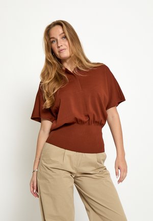 Frau mit langen blonden Haaren, die ein rostfarbenes Kurzarm-Top und beige Hosen trägt, steht vor einem schlichten hellen Hintergrund.