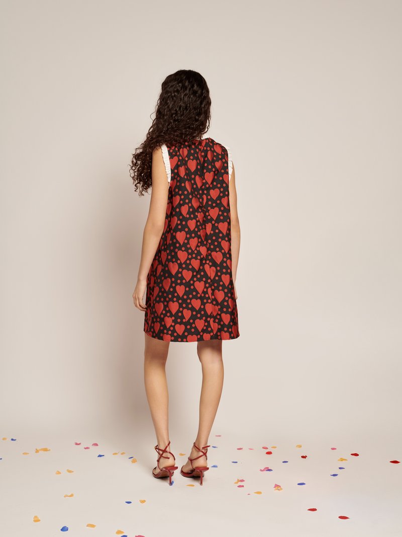 Vestido preto sem mangas com um padrão de corações vermelhos, apresentando uma silhueta rodada. Modelo usa saltos vermelhos com tiras, rodeada por confete colorido.