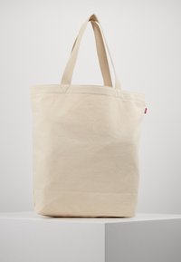 Beige canvas toteväska med dubbla tyghandtag, rektangulär form och sydda detaljer. Har ett litet rött logotypmärke.