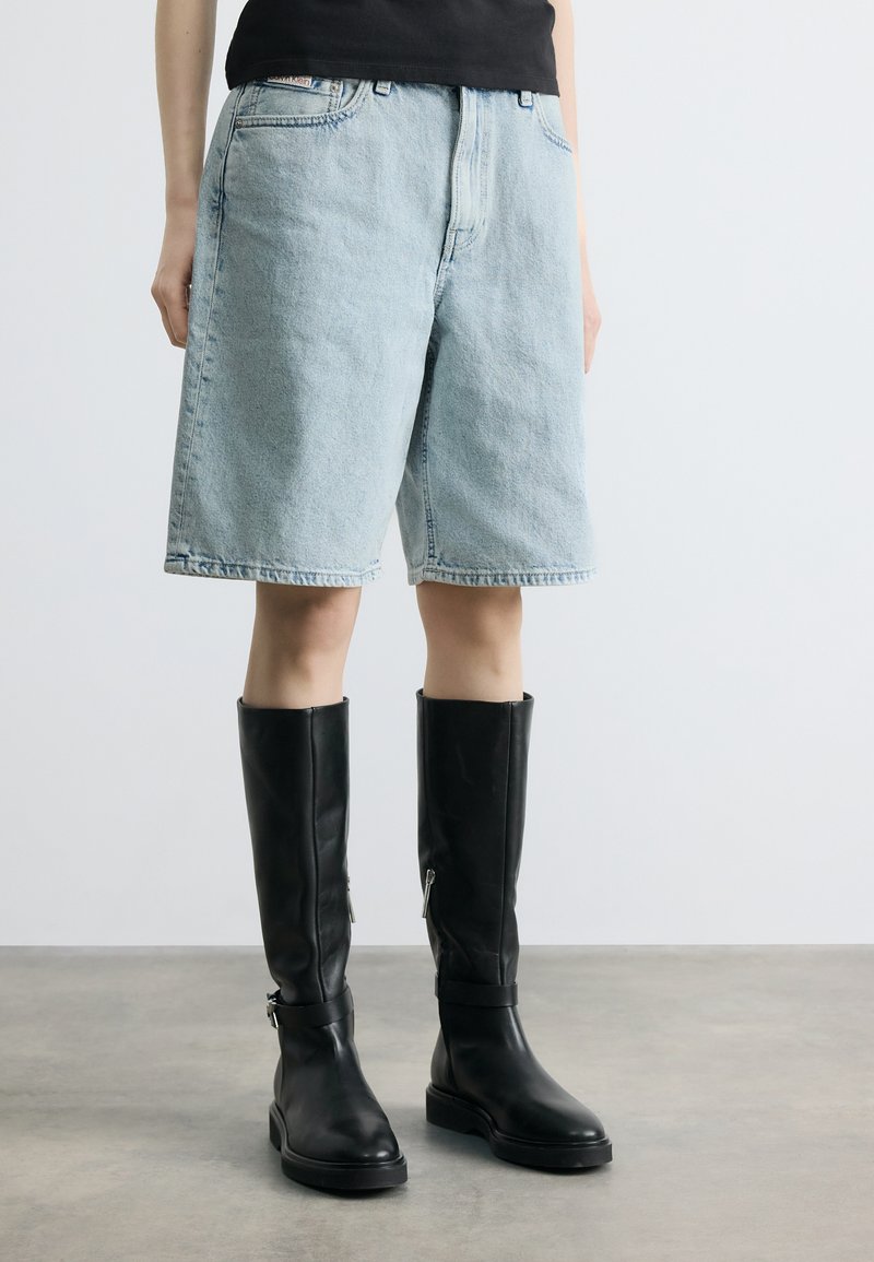 Άτομο που φοράει ανοιχτό μπλε τζιν σορτς και μαύρες δερμάτινες μπότες μέχρι το γόνατο, στέκεται σε γκρι δάπεδο με φόντο έναν απλό λευκό τοίχο.