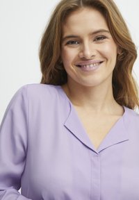 Lavendelfarbene Bluse aus glattem Stoff mit einem ausgestellten Kragen und subtilen Knopfdetails, die eine lockere Passform und eine leichte Textur bietet.