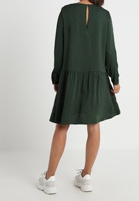 Vestido verde con lunares, de manga larga, con dobladillo rizado y un escote en forma de lágrima en la espalda, combinado con zapatillas blancas.
