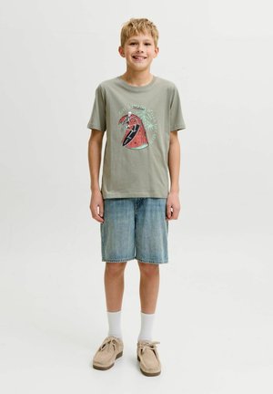 Smilende dreng iført lysegrå grafisk t-shirt, denimshorts, hvide sokker og beige sko, der står foran en simpel hvid baggrund.