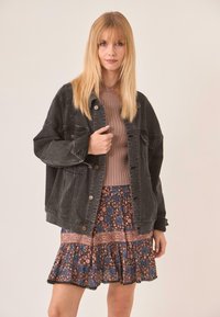 Chaqueta de mezclilla negra con un corte relajado, que presenta grandes botones y bolsillos, combinada con una blusa marrón de canalé y una falda con estampado floral.