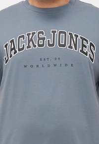 Grå bomulls t-skjorte med rund hals og fet svart tekst som sier "JACK&JONES" med "EST. 90 WORLDWIDE" nedenfor.
