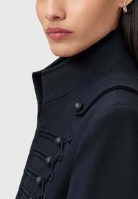 Granatowy płaszcz wełniany z wysokim kołnierzem, z ozdobnymi czarnymi guzikami i plecionymi akcentami na froncie. Zbliżenie na teksturę tkaniny.