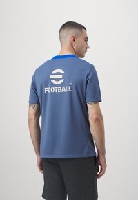 Camisa de fútbol gris con cuello azul, que presenta un gran logo blanco en la parte posterior. Confeccionada con tejido transpirable y de manga corta.