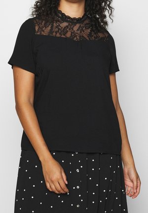 Femme portant un haut noir à manches courtes avec un empiècement en dentelle et une jupe noire à pois blancs avec des boutons sur le devant, debout devant un fond neutre.