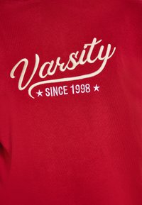 Felpa rossa con testo ricamato bianco: "Varsity" in stile corsivo e "SINCE 1998" in carattere dritto, con accenti a forma di stella.