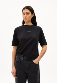 Schwarzes Baumwoll-T-Shirt mit kurzen Ärmeln, runder Halslinie und weißem Logoschriftzug auf der Brust. Kombiniert mit hochgeschlossenen schwarzen Jeans.