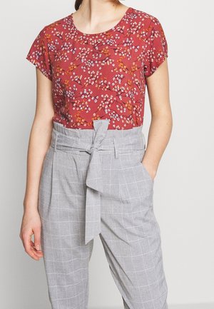 Femme portant un chemisier floral à manches courtes couleur rouille, associé à un pantalon taille haute gris clair à carreaux, ceinturé avec une ceinture en tissu.