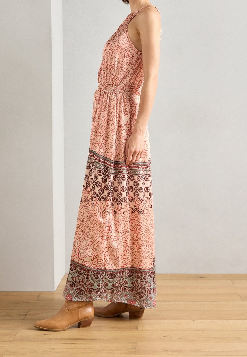Femme debout à l'intérieur, portant une robe maxi sans manches à motifs et des bottines marron clair sur un sol en bois contre un mur uni.