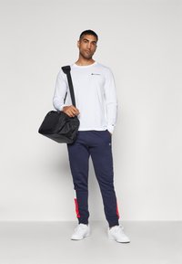 Hvitt langermet skjorte med logo, marineblå joggebukser med røde sideakenter, og hvite sneakers; person som holder en svart skulderveske.