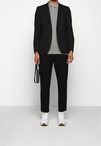 Schwarze Blazer über einem grauen Polo-Shirt, kombiniert mit schwarzen, taillierten Hosen und weißen Sneakers. Hält in einer Hand einen schwarzen Gurt.