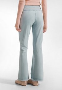 Mujer con pantalones anchos azul claro, bailarinas beige y una blusa de manga larga color durazno, posando frente a un fondo blanco liso.