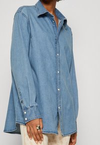 Persona che indossa una camicia di jeans leggera blu chiaro con maniche lunghe, abbinata a pantaloni beige con motivi e un anello metallico spesso sulla mano sinistra.