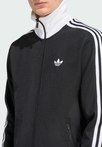 Μαύρο αθλητικό μπουφάν με ψηλό γιακά, άσπρο φερμουάρ και τρεις άσπρες ρίγες σε κάθε μανίκι. Διαθέτει ένα μικρό άσπρο λογότυπο στο στήθος.