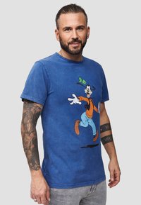 Sininen puuvillapaita, jossa on graafinen printti sarjakuvahahmosta, joka on pukeutunut oranssiin ja siniseen asusteeseen. Paidassa on lyhyet hihat ja pyöreä kaula-aukko.