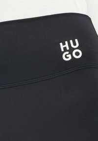 Svarta sportleggings med en slät textur och sömmar. Har en vit broderad "HUGO"-logotyp på midjebandet.