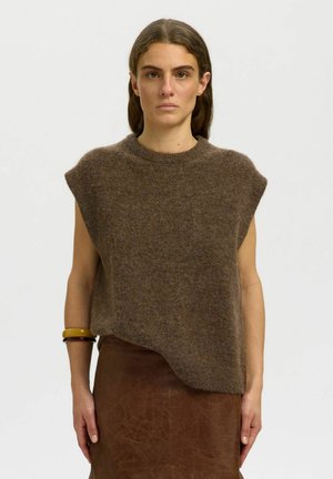 Kvinde med glat mørkt hår iført ærmeløs, fluffy brun sweater og brun lædernederdel, stående mod en ensfarvet lys baggrund.