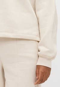 Beige pulóver és hozzá illő nadrág, puha szövetből készült. Részletes bordás mandzsettákkal és szegéllyel rendelkezik, finom varrási részletekkel. Sima tapintású.