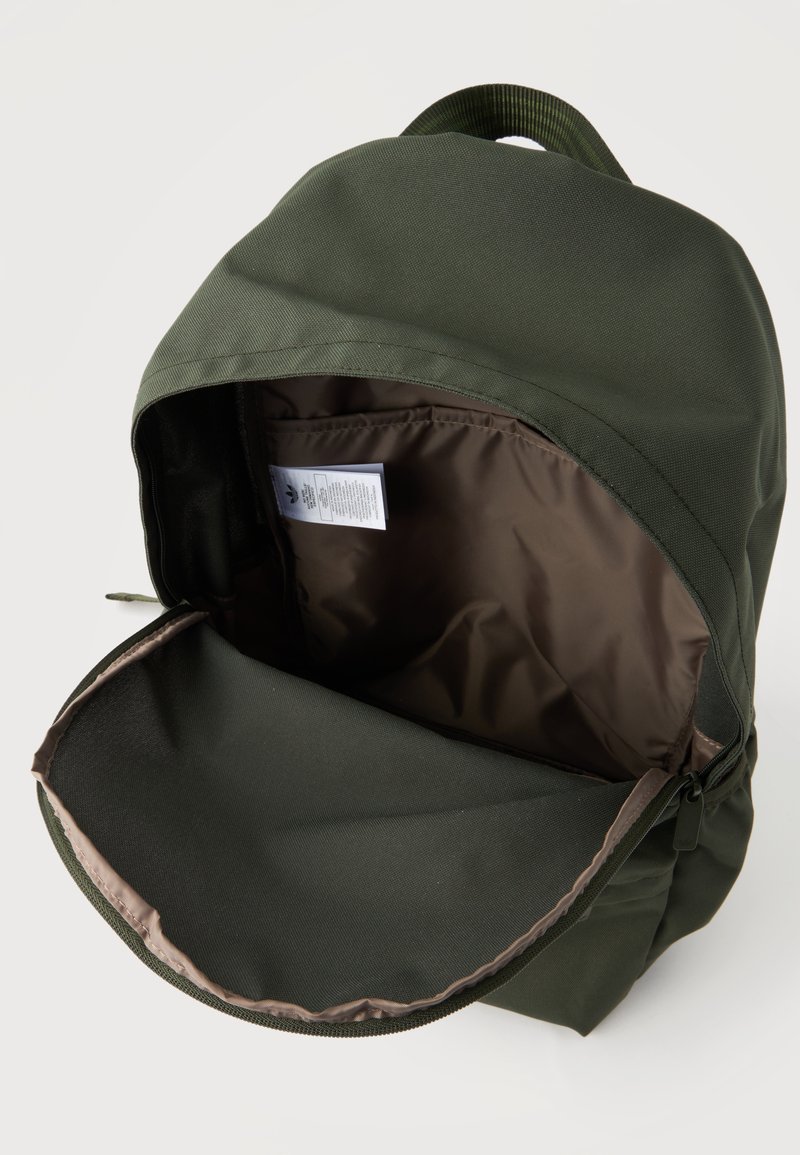 Sac à dos vert foncé avec le compartiment principal ouvert, montrant un intérieur vide doublé de brun et une étiquette d'entretien visible à l'intérieur.
