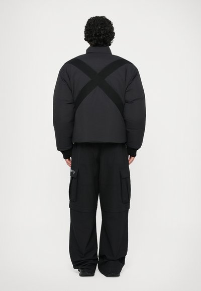 Doudoune noire avec un design court exagéré, présentant un détail de sangle en forme de X dans le dos. Assortie à un pantalon cargo noir à jambes larges.
