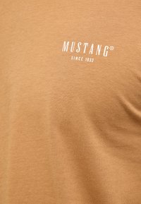 T-shirt Mustang en coton marron clair, avec un logo blanc "MUSTANG SINCE 1932" imprimé sur le devant, texture lisse, col rond.