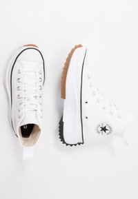 Białe sneakersy typu high-top z teksturowanej kanwy, gumowym czubkiem i poszarpaną, w kolorze gumy, podeszwą. Posiadają naszywkę z logo z boku.
