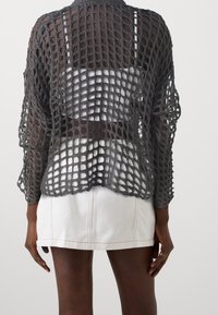 Cárdigan gris de punto abierto con mangas largas, que presenta un diseño holgado y aireado. Llevado sobre una blusa blanca y combinado con una falda de mezclilla blanca.