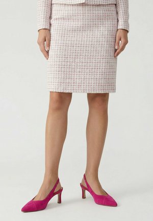 Vrouw draagt een lichtroze tweed rokkenpak en felroze suède slingbackhakken, staand tegen een effen witte achtergrond.