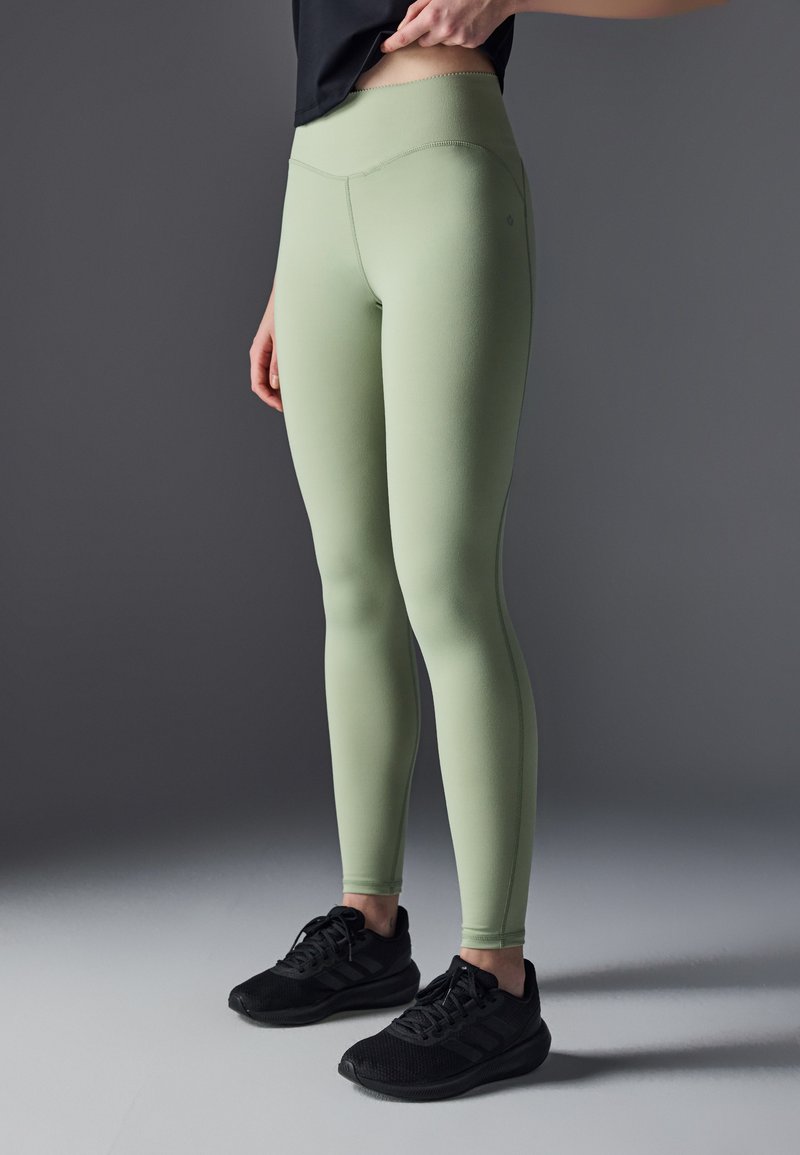 Leggings de color verde claro, de talle alto y con una textura suave. Combinados con zapatillas deportivas negras, que cuentan con un diseño de cordones y paneles de malla.