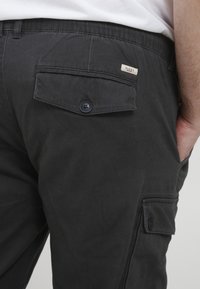 Schwarze Cargo-Hosen mit einer Gesäßtasche mit Klappe und Knopfverschluss, aus robustem Stoff mit glatter Textur hergestellt.